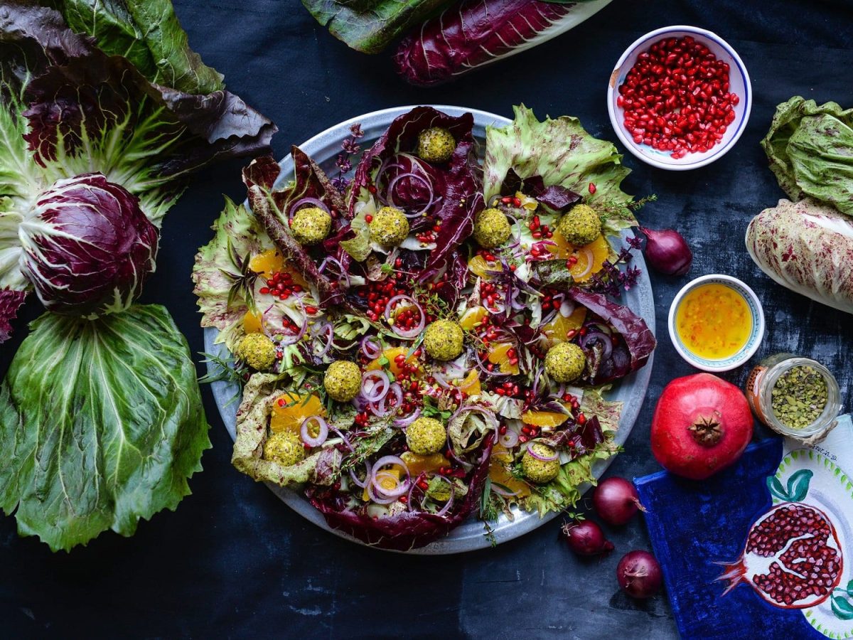 Radicchio mit Pistazien-Ricotta-Bällchen, Orangen und Granatapfelkernen in der Draufsicht, dunkelblauer Untergrund, daneben viele zutaten wie Salat und Granatapfel.