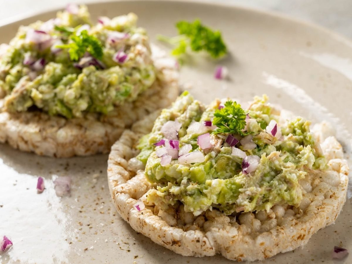 Zwei Reiswaffeln mit dem Avocado-Thunfischsalat auf einem weißen Teller und hellem Untergrund.