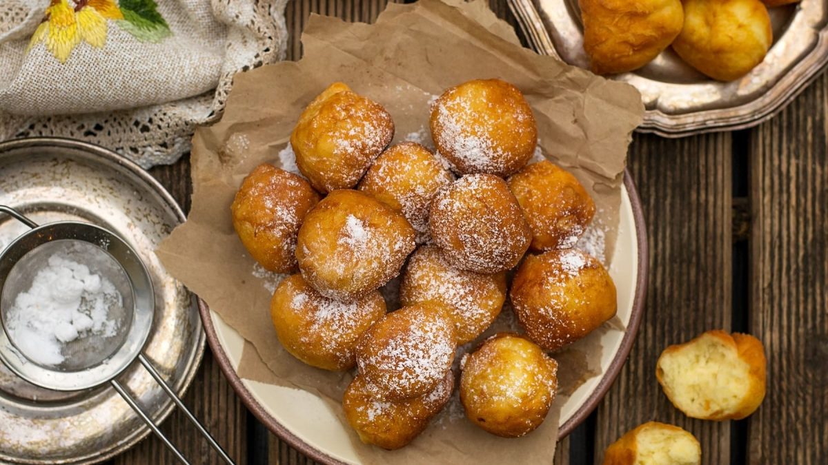 Mehrere der frittierten Ricotta-Bällchen mit Zitrone auf einem rustikalen Holztisch mit Puderzucker und Tischdecke. Direkt von oben fotografiert.