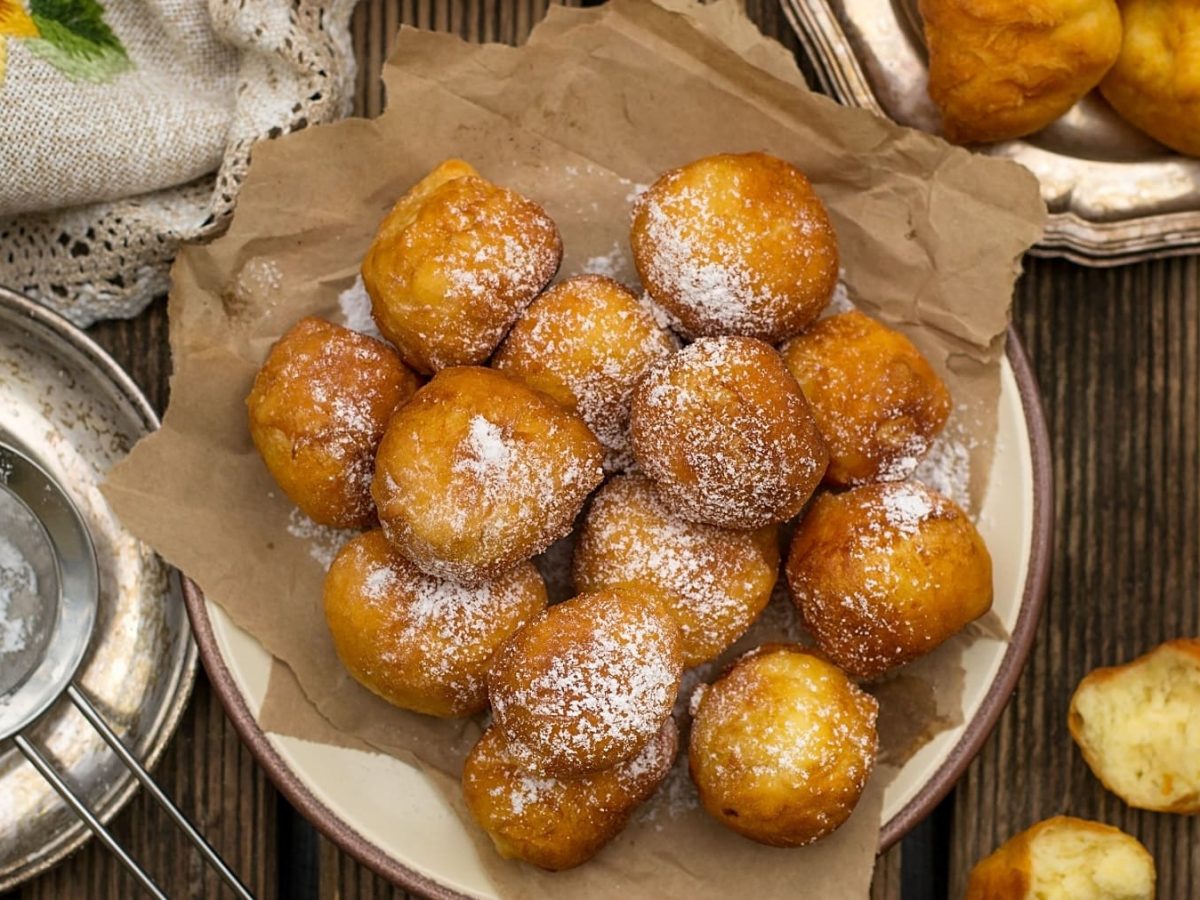 Mehrere der frittierten Ricotta-Bällchen mit Zitrone auf einem rustikalen Holztisch mit Puderzucker und Tischdecke. Direkt von oben fotografiert.