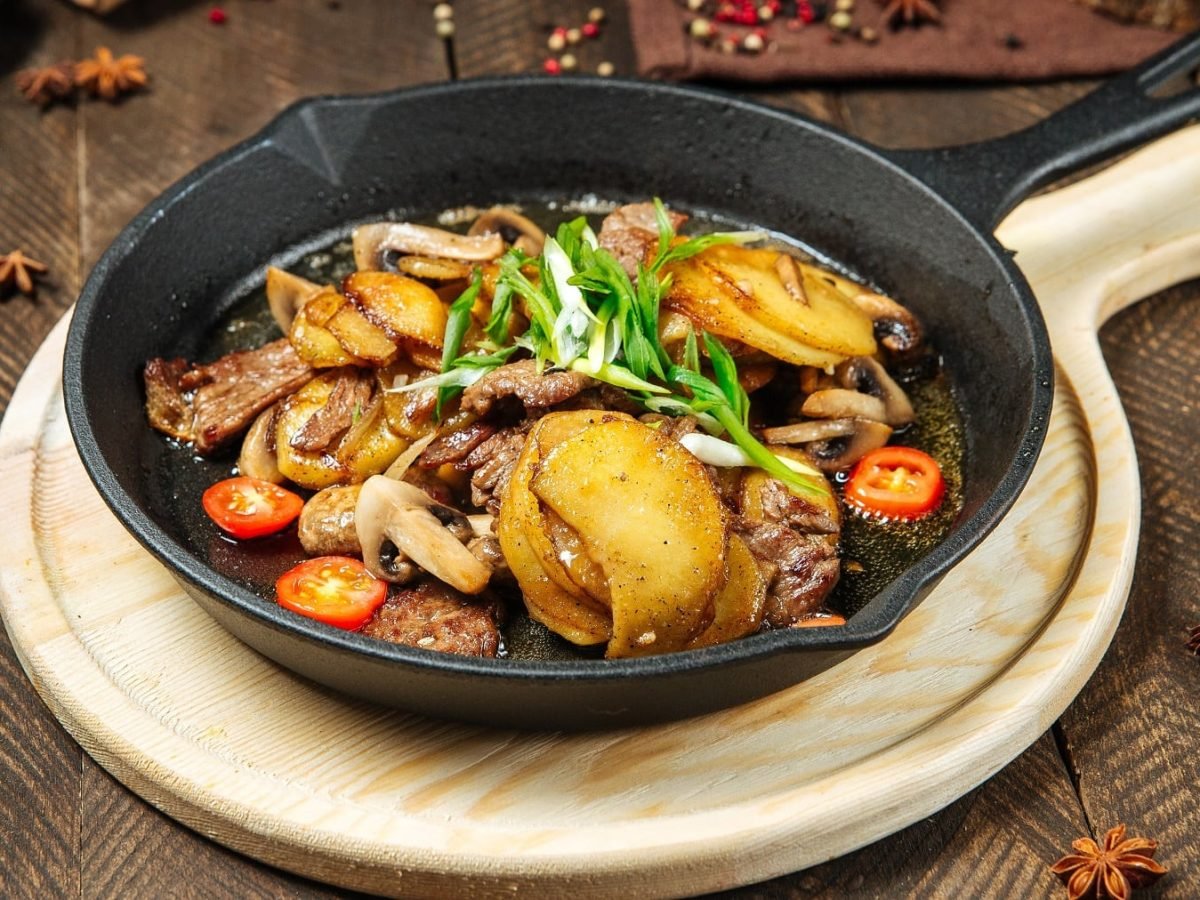 Eine gusseiserne Pfanne auf einem Holzbrett mit dem Rindfleisch-Kartoffelgericht auf dunklem Holzuntergrund mit Gewürzen. Seitlich von leicht schräg oben fotografiert.