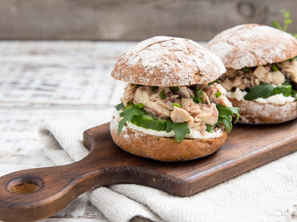 Zwei Roggenburger mit Hüttenkäse und Thunfisch Burger zum Frühstück auf Holzbrett mit Griff auf Tuch und hellem Untergrund. Im Bild Rucola. Frontalansicht.