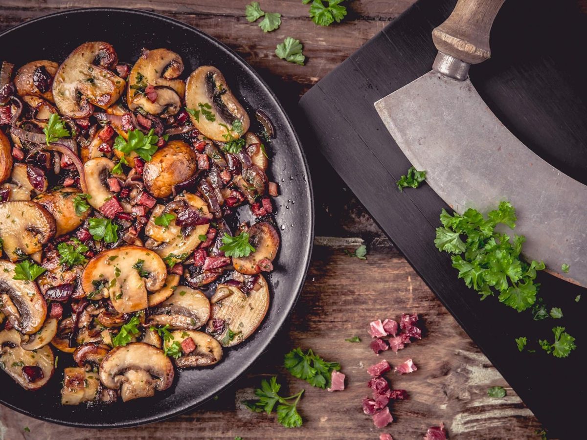 Draufsicht: eine Schinken-Champignon-Pfanne liegt auf einem Holztisch. Daneben liegen frisch gehackte Peterslilie und ein Wiegemesser,