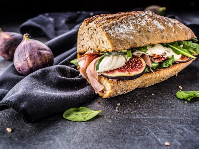 Frontal: Ein Schinken-Feigen-Sandwich mit Mozzarella auf einem Tuch, das auf dunklem Untergrund liegt. Drum herum liegen frische Feigen und Salatblätter.