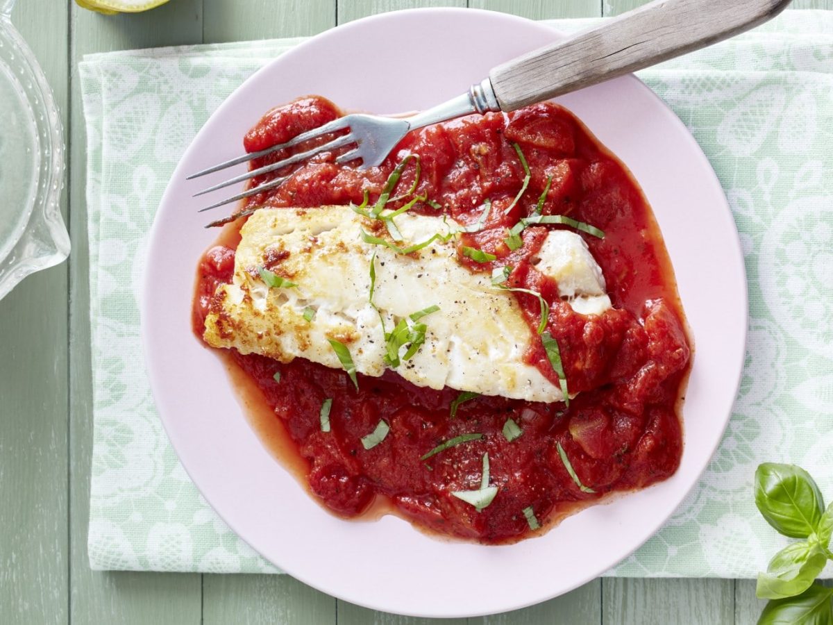 Ein rosafarbener Teller mit Seelachs mit Tomatensugo auf einem hellgrünen Tisch.