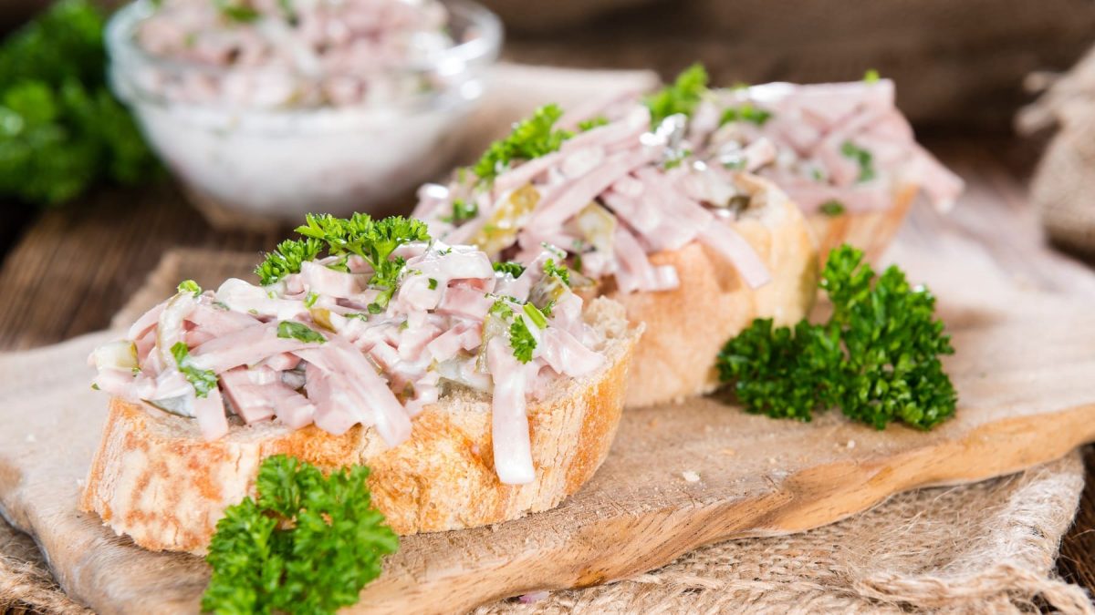 Ein Holzbrett mit zwei Weißbrotscheiben und dem Fleischsalat sowie frische Petersilie. Alles auf einem Holztisch.