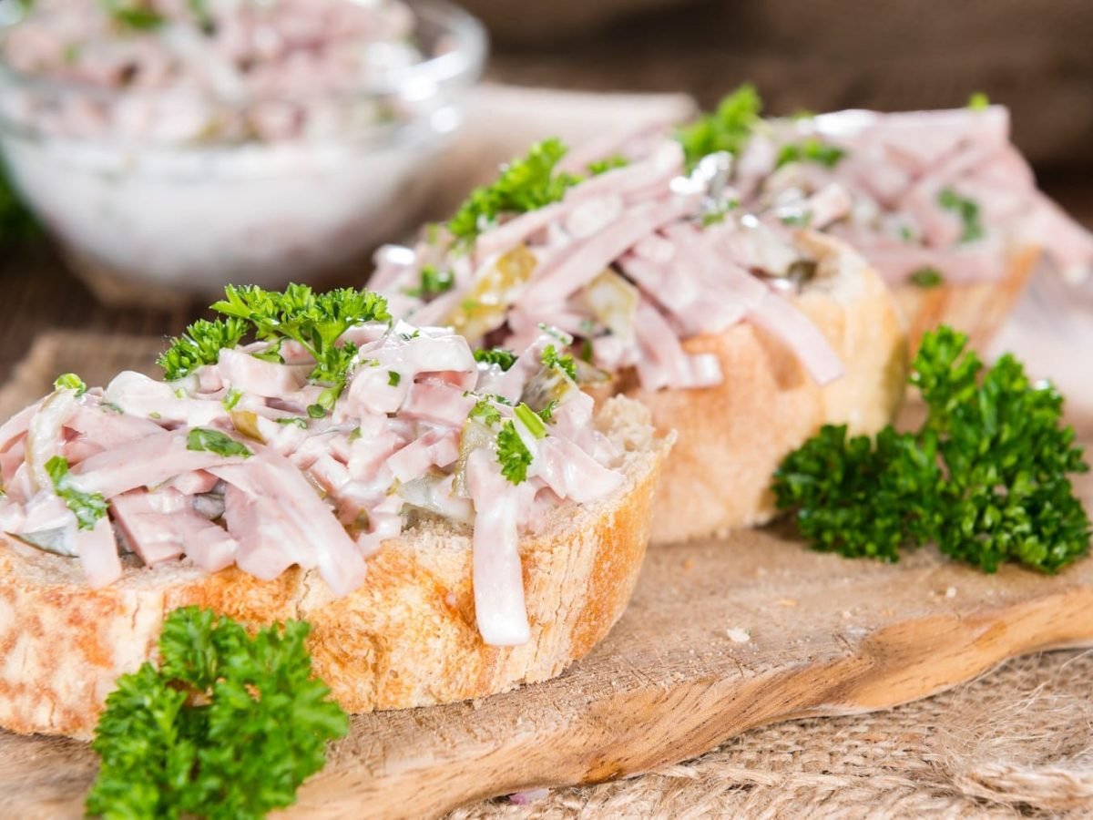 Ein Holzbrett mit zwei Weißbrotscheiben und dem Fleischsalat sowie frische Petersilie. Alles auf einem Holztisch.