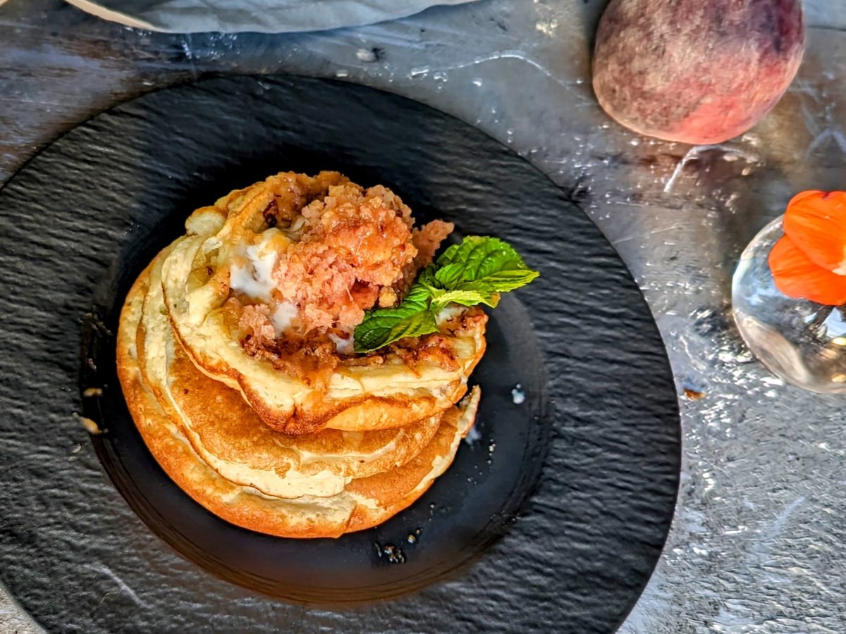 Ein schwarzer Teller mit Pancakes und geriebenem, gefrorenem Pfirsich auf grauem Steinuntergrund. Daneben eine orangene Blüte, ein Pfirsich sowie eine graue Tischdecke.