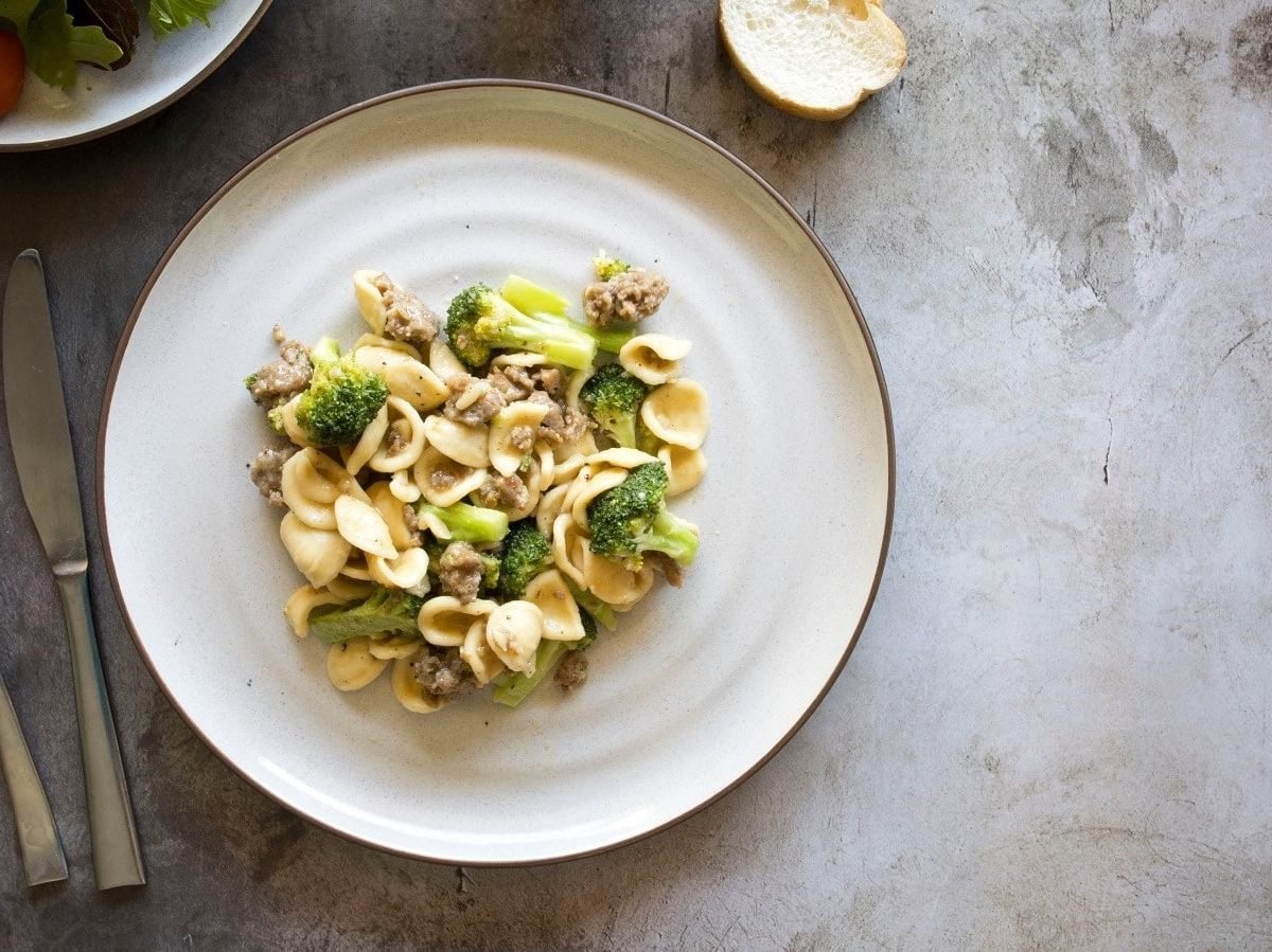 Ein weißer Teller mit Orecchiette mit Salsiccia und Brokkoli auf einem grauen Tisch.