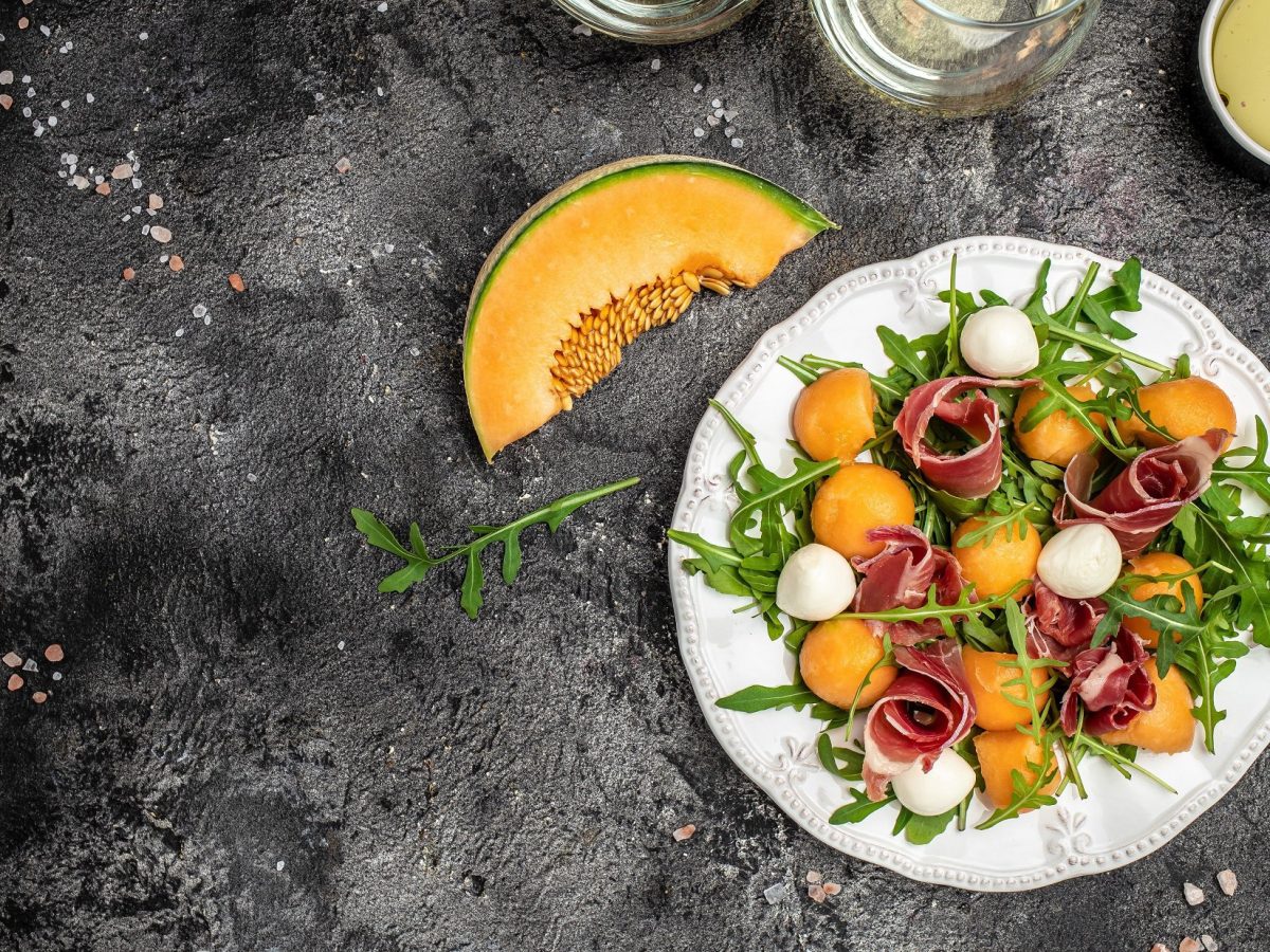 Draufsicht: Ein heller Teller süßer und fruchtiger Sommersalat mit Cantaloupe-Melone auf dunklem Untergrund.