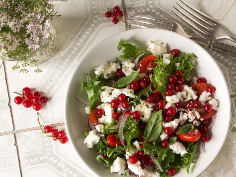 Draufsicht: Ein rot-weiß-kariertes Küchenhandtuch auf dem eine weiße Schüssel mit einem Sommersalat mit Johannisbeeren und Feta steht. Daneben ist einen Blumenstraß mit Holler zu sehen.