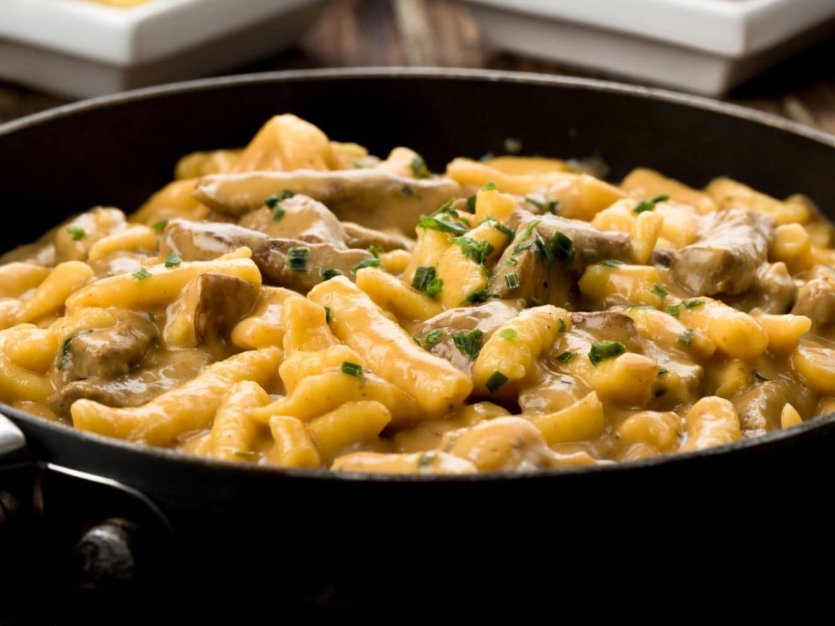 Spätzlepfanne mit Steinpilzen und Steakstreifen aus Flanksteak in einer schwarzen Pfanne auf Holztisch. Im Hintergrund angedeutet zwei weiße Schalen. Draufsicht.