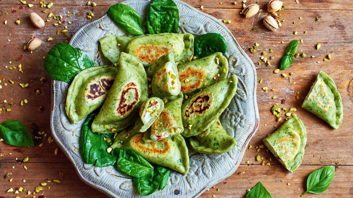 Draufsicht: Eine Teller selbst gemachte Spinat-Ravioli mit Ricotta-Pistazien-Füllung sind auf einem orientalisch angehauchtem Teller. Drum herum liegen Pistazienkerne und Spinatblätter.