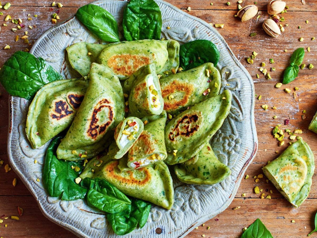 Draufsicht: Eine Teller selbst gemachte Spinat-Ravioli mit Ricotta-Pistazien-Füllung sind auf einem orientalisch angehauchtem Teller. Drum herum liegen Pistazienkerne und Spinatblätter.