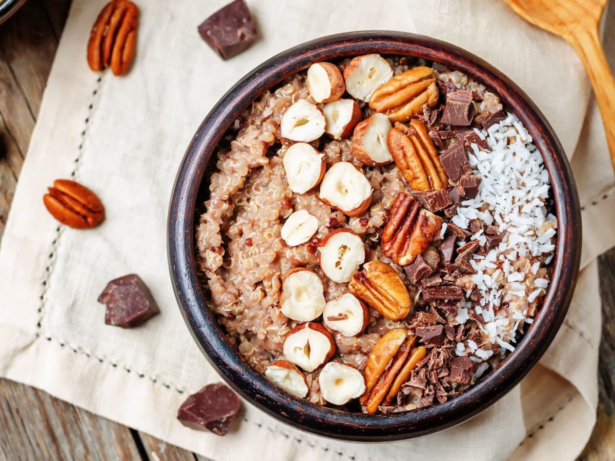 Eine Schüssel süßer Quinoa mit Nüssen, Schokolade und Kokosraspeln in der Draufsicht, daneben ein Holzlöffel und Zutaten.