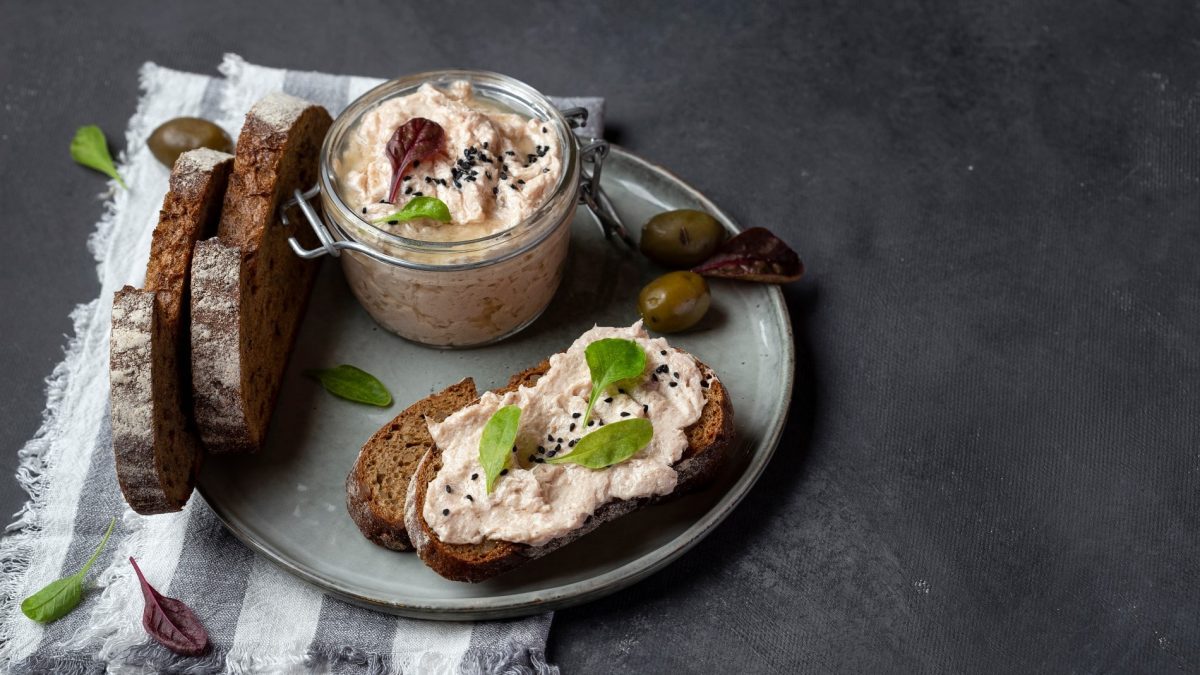 Ein grauer Teller mit veganer Thunfisch-Mayo in einem Bügelverschlussglas und Schwarzbrotscheiben. Ein Brot davon ist bestrichen mit veganer Thunfisch-Mayo und garniert mit Basilikum und schwarzem Sesam. Der Hintergrund ist grau und der Teller steht auf einem weiß grau gestreiften Geschirrtuch.