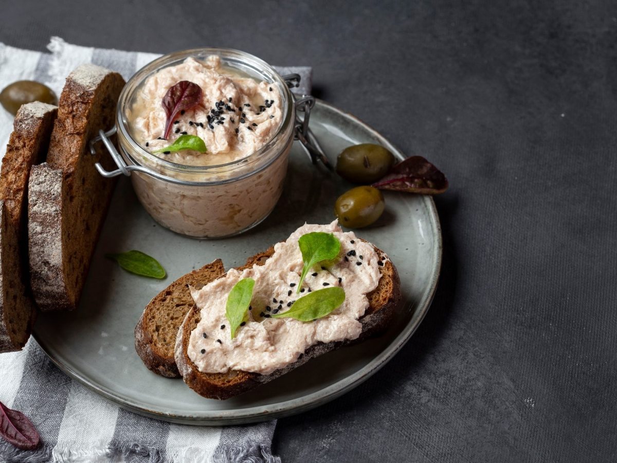 Ein grauer Teller mit veganer Thunfisch-Mayo in einem Bügelverschlussglas und Schwarzbrotscheiben. Ein Brot davon ist bestrichen mit veganer Thunfisch-Mayo und garniert mit Basilikum und schwarzem Sesam. Der Hintergrund ist grau und der Teller steht auf einem weiß grau gestreiften Geschirrtuch.