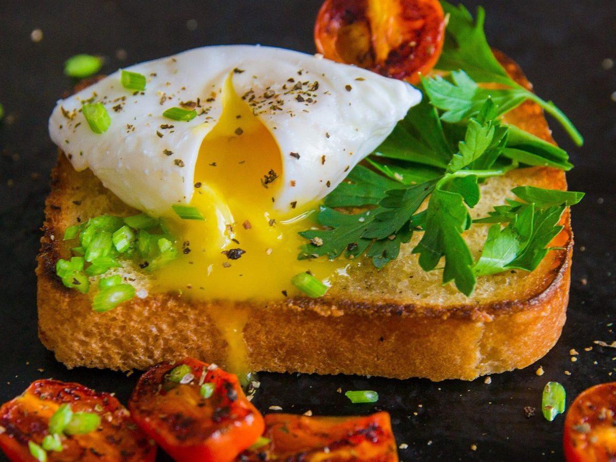 Frontalsicht: Auf einem dunklen Untergrund liegt ein Toast mit pochiertem Ei und gebratenen Tomaten und frischen Kräutern. Die Tomaten liegen um das Toast herum.