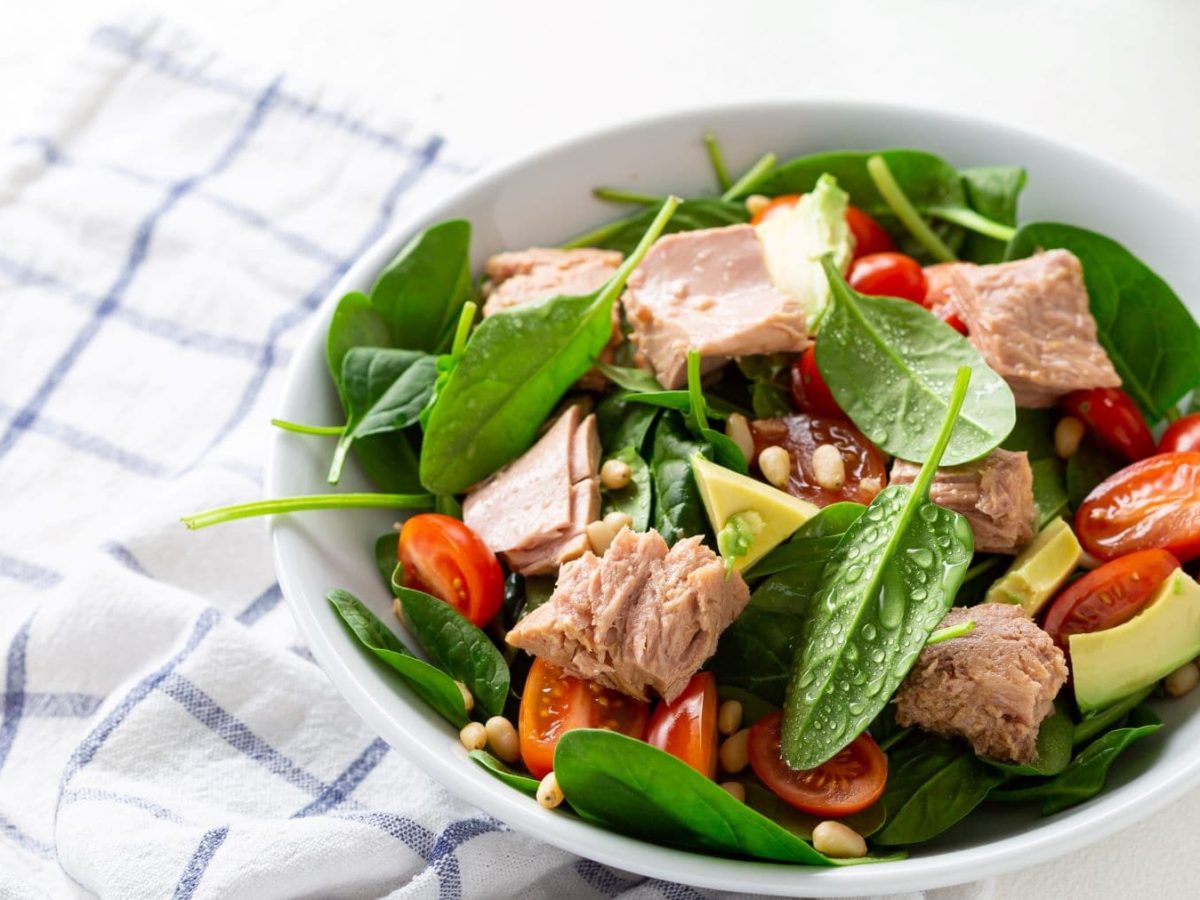 Frischer, schneller Tomaten-Spinat-Salat mit Thunfisch, Avocado und Pinienkerne in weißer Salatschüssel auf Tuch und hellem Untergrund. Draufsicht.