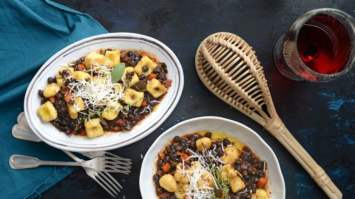 Zwei Teller Topini mit Kichererbsenragout in der Draufsicht, daneben eine Schöpfkelle aus Holz und ein Glas Rotwein.