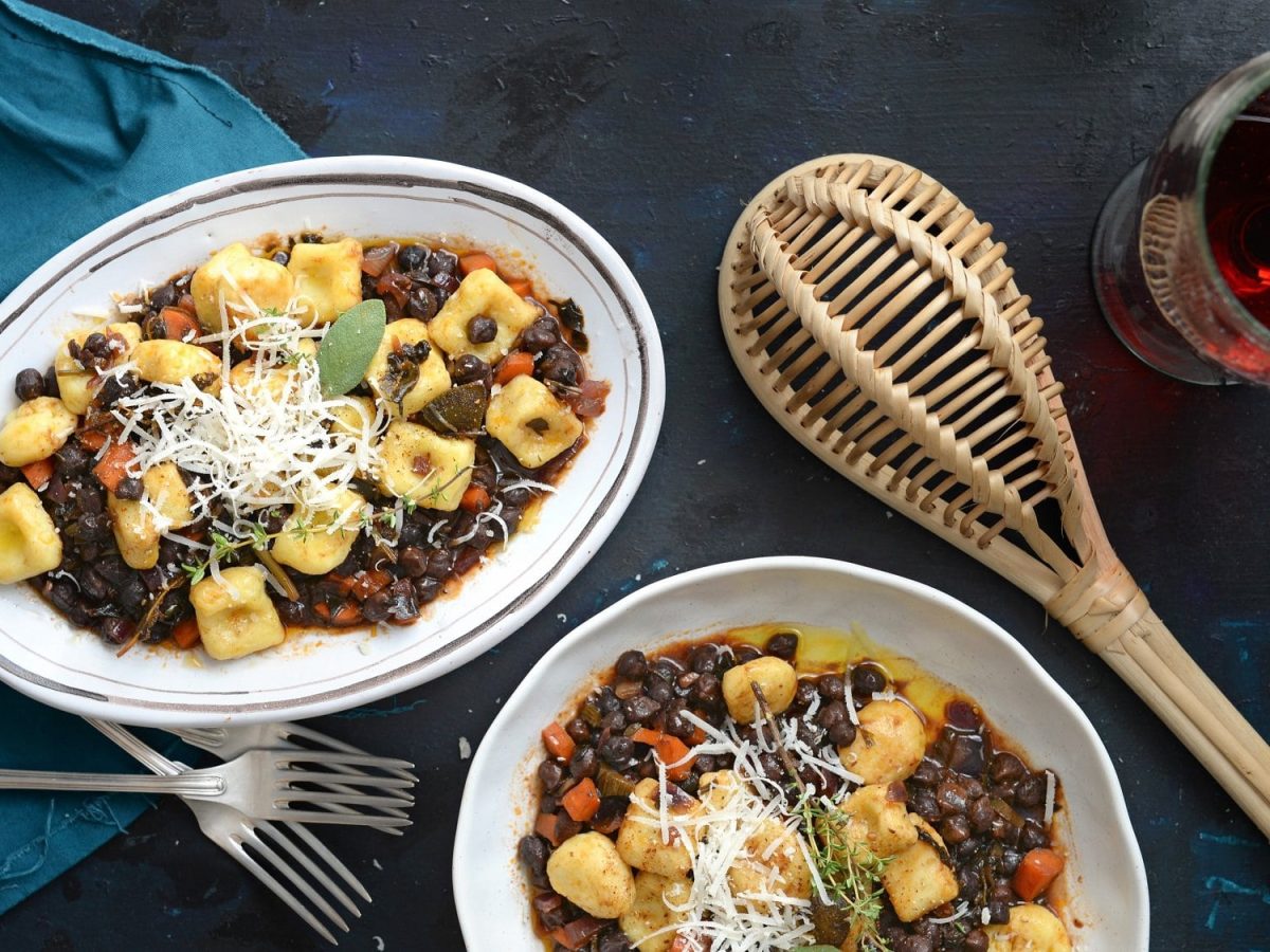 Zwei Teller Topini mit Kichererbsenragout in der Draufsicht, daneben eine Schöpfkelle aus Holz und ein Glas Rotwein.