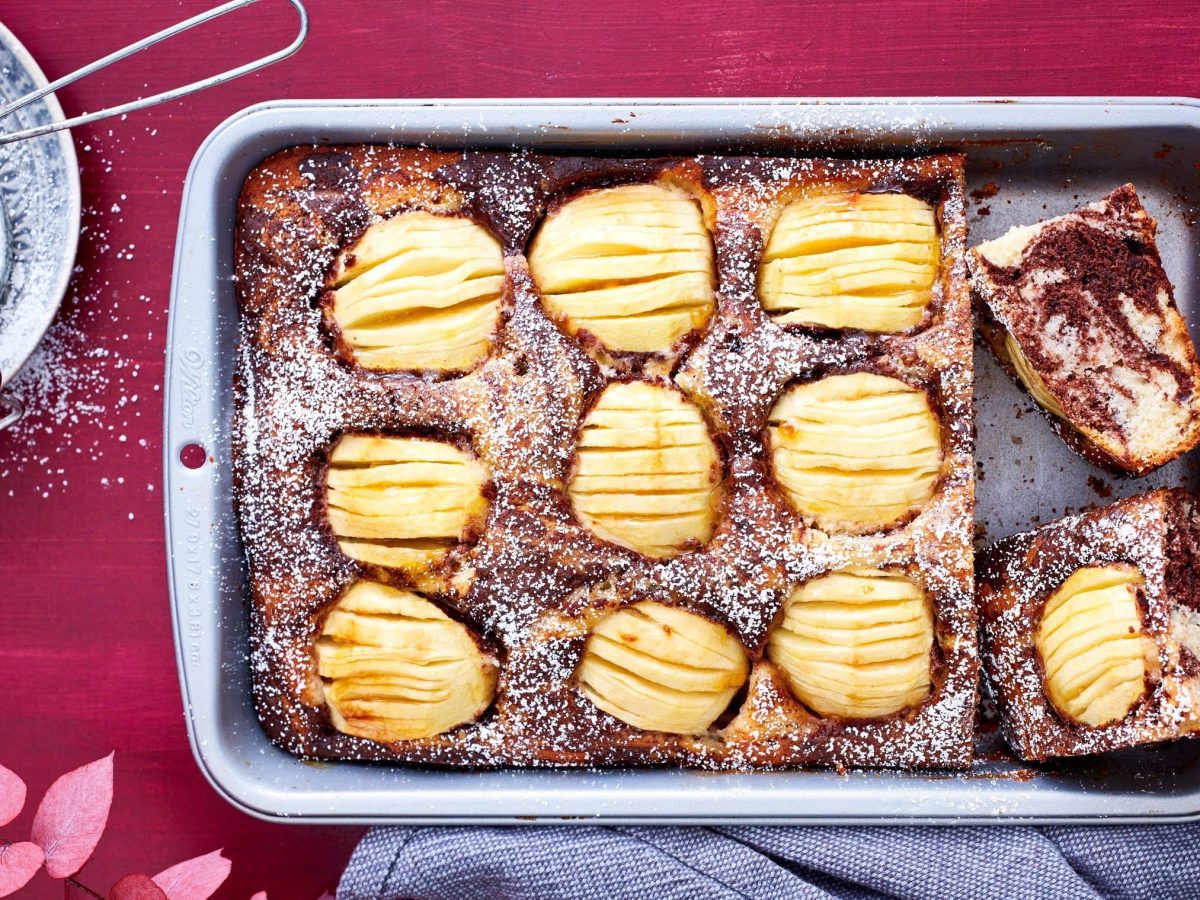 In einer Ofenform ist ein versunkener Apfel-Marmorkuchen gebacken. Er ist mit Puderzucker bestreut worden.