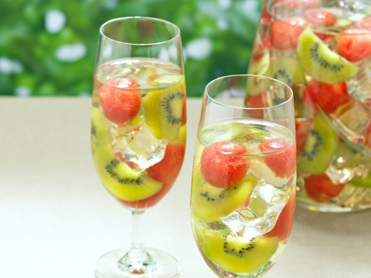 White Sangria mit Melone und Kiwi in einer Schale und zwei gefüllte Sektgläser auh hellem Tisch. Außenaufnahme. Frontalansicht.