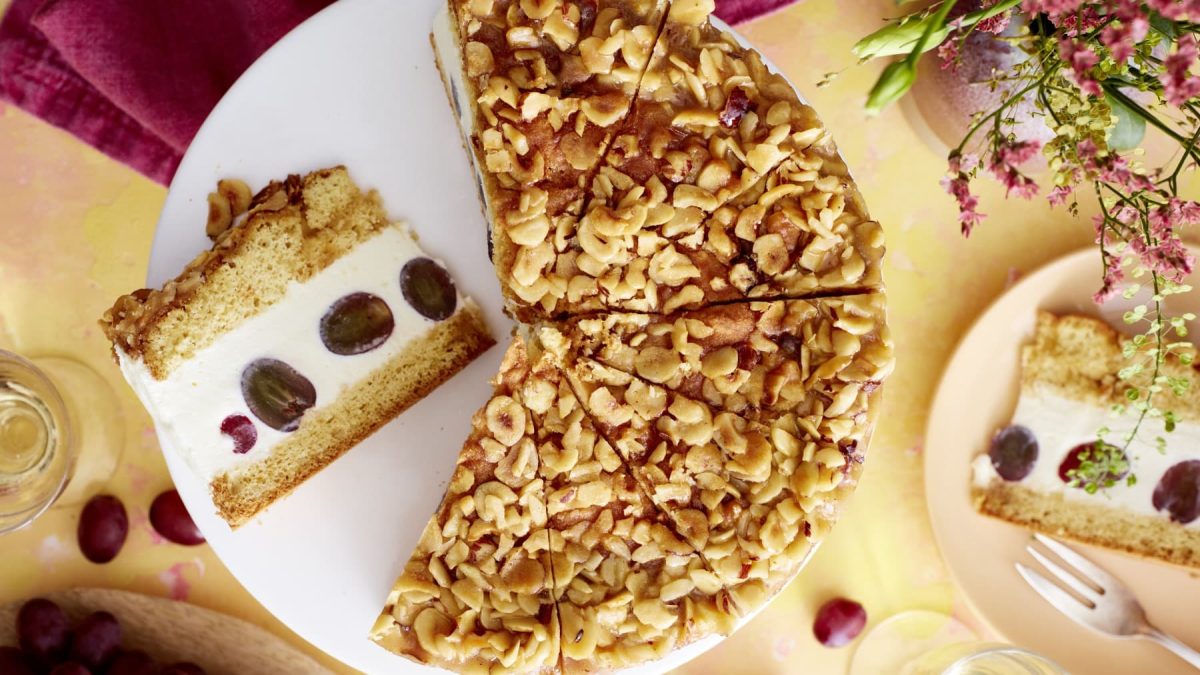 Eine Weißwein-Trauben-Torte angeschnitten auf einem weißen Servierteller auf einem gelben Hintergrund. Oben auf dem Foto ein rotes Geschirrtuch und Blumen, links unten rote Trauben und rechts ein Teller mit einem Stück Torte.