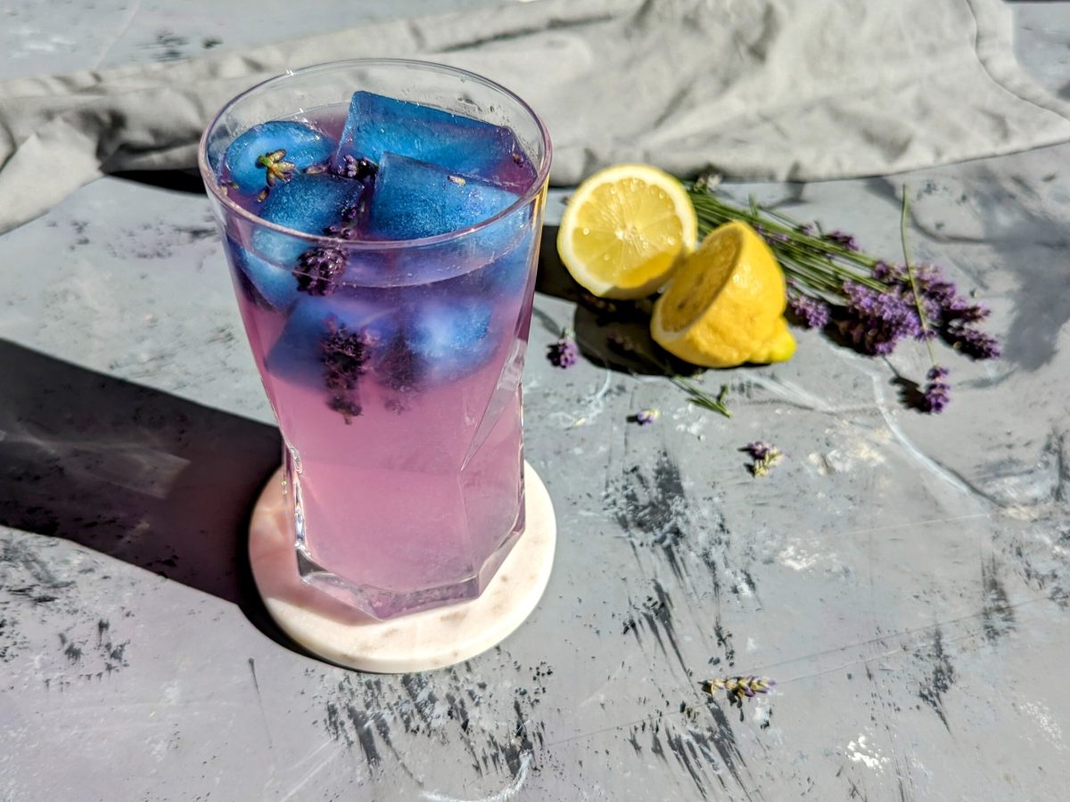 Ein Glas mit der Zitronenlimonade und den blauen Eiswürfeln auf einem Steintisch. Die Limonade hat sich schon leicht pink verfärbt. Im Hintergrund Zitrone und Lavendel.