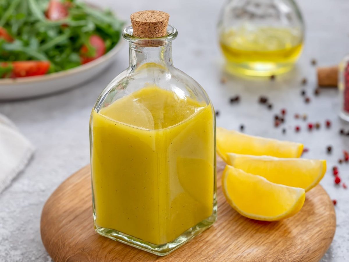 Eine Glasflasche mit dem Zitronendressing auf einem hellen Tisch mit rotem Pfeffer und einem Teller mit Salat.