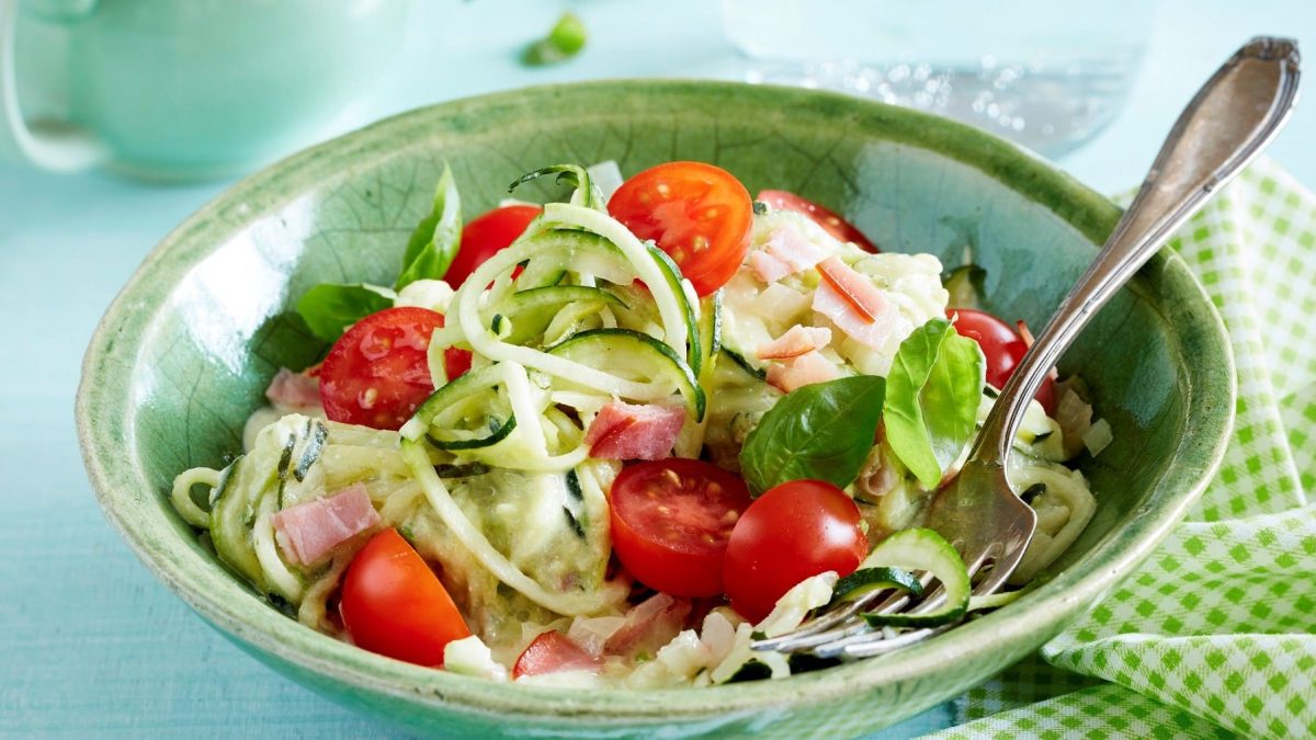 In einer Schüssel ist eine Portion Zucchini-Spaghetti angerichtet. Sie ist mit Tomaten und Basilikum verfeinert.