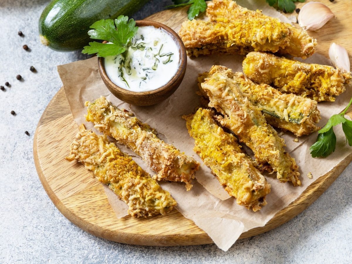 Ein Holzbrett mit den Zucchini-Sticks auf grauem Untergrund mit Zucchini, Knoblauch und frischer Petersilie.