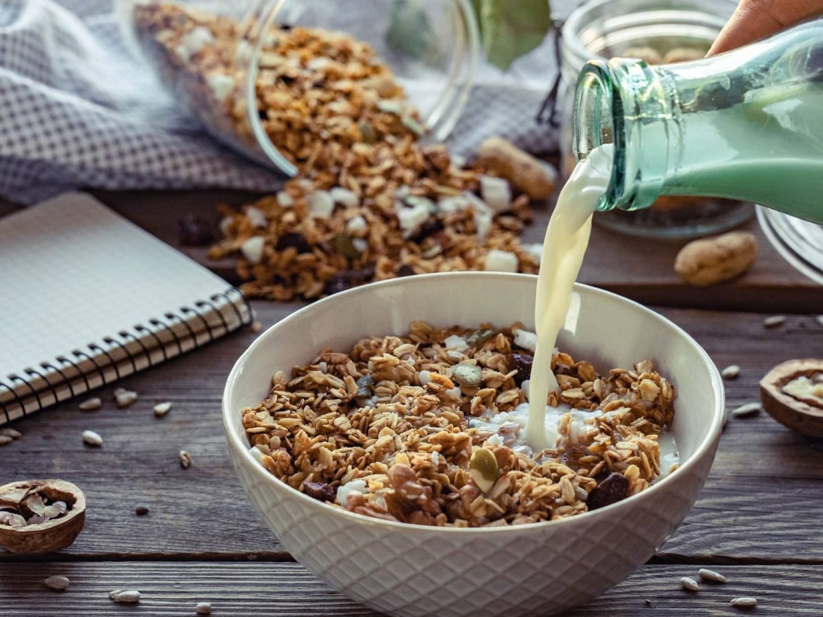 Frontalsicht: Eine Schüssel mit Apfel-Nuss-Granola steht auf einem Tisch. Eine weibliche Hand hält eine Glasflasche Milch in der Hand und schüttet Milch in die Schahle. Drum herum liegen ein Küchenhandtuch und ein Schreibblock.