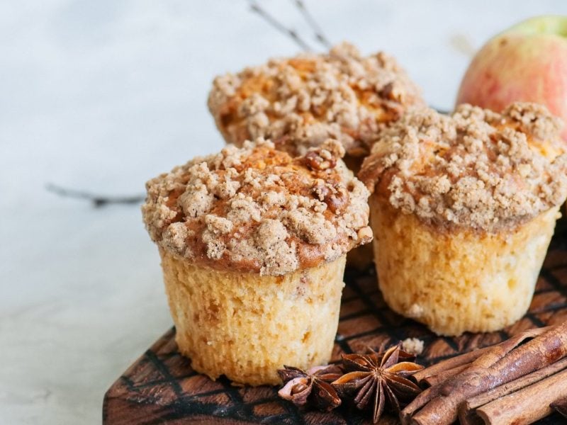 3 Apfel-Streusel-Muffins auf einem Brett, auf dem auch Zimtstangen, angeschnittene Äpfel und Anis liegen.