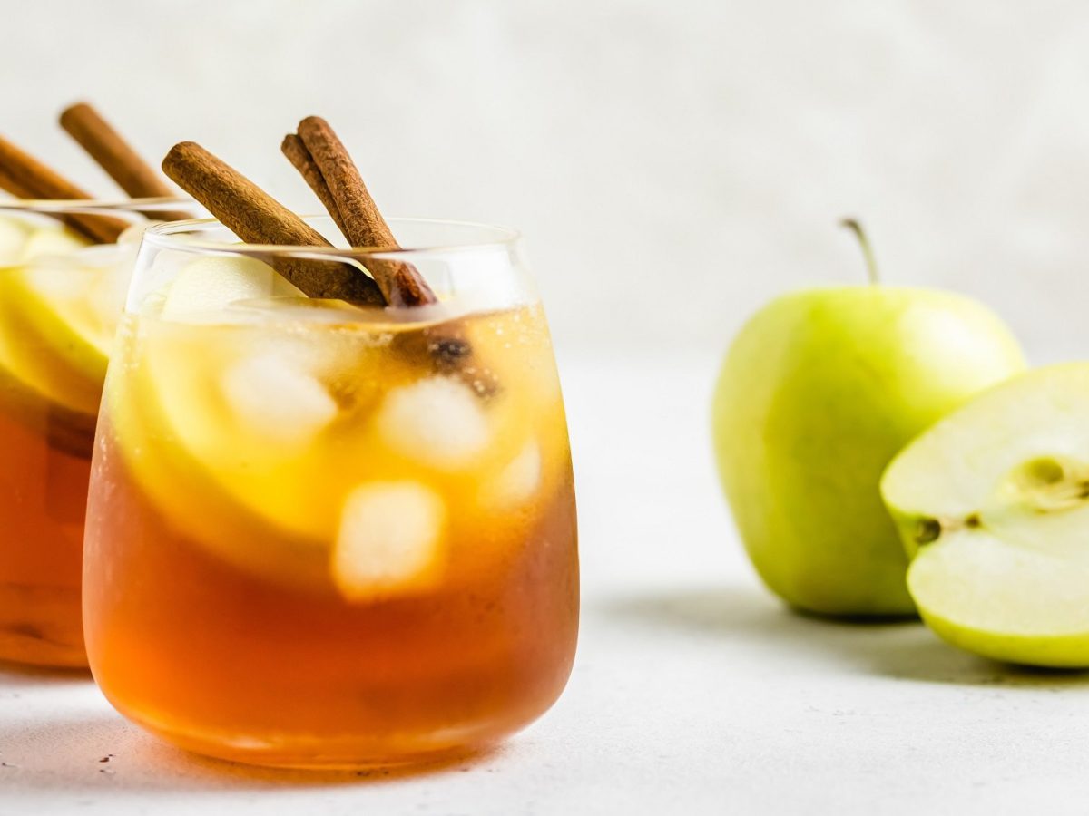 Zwei Gläser Apple Cider Spritz mit Eiswürfeln, Apfelscheiben und Zimtstangen garniert, daneben liegen ein ganzer und ein angeschnittener Apfel.