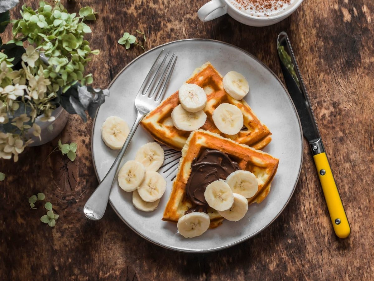 Ein Teller mit 2 Bananenbrot-Waffeln, frischen Bananenscheiben und Schokocreme in der Draufsicht, daneben Besteck und eine Tassee Cappuccino sowie Blumen.