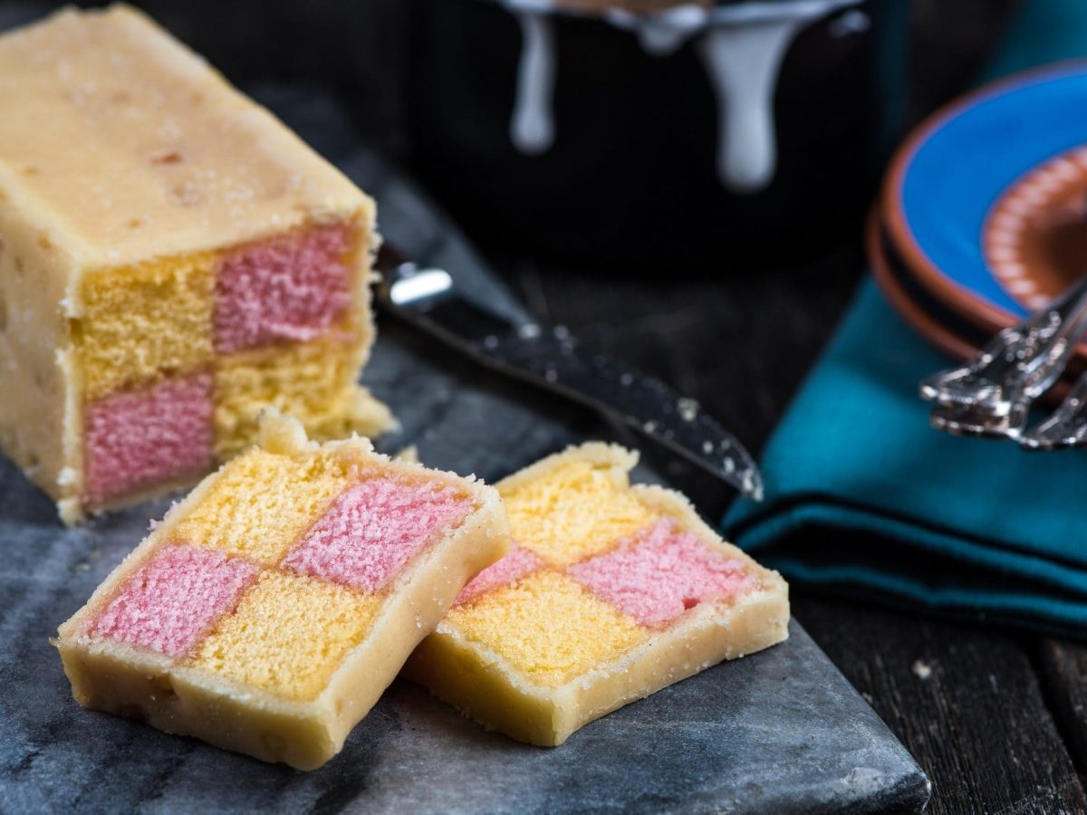 Draufsichtschräg: ein angeschnittener Battenberg Cake auf einem dunklen Brettchen auf dunklem Untergrund. Der Kuchen hat innen zwei verschiedene Farben, die Karo-Förmig angeordnet sind. Es handelt sich um die Farben gelb und rosa.