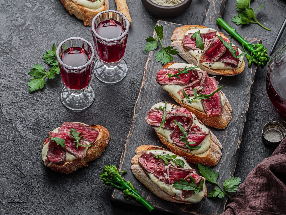 Draufsicht: Dunkler Untergrund und ein Holzbrettchen mit mehreren Beef-Bruschette mit Avocado-Creme. Daneben stehen eine Karaffe und eine Glaskaraffe mit Rotwein.