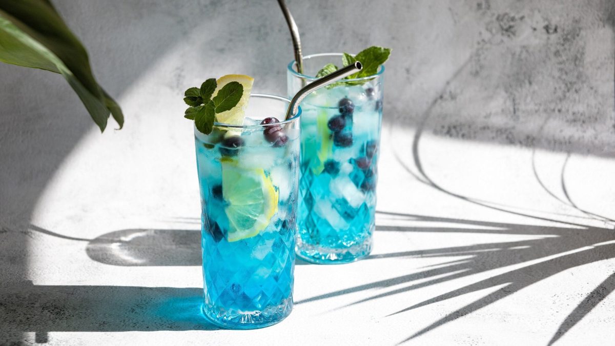 2 Gläser Blue Lagoon mit Eis, Blaubeeren, einer Zitronenscheibe und Metallstrohhalm und Minze garniert in hellem Setting, man sieht Palmenschatten.