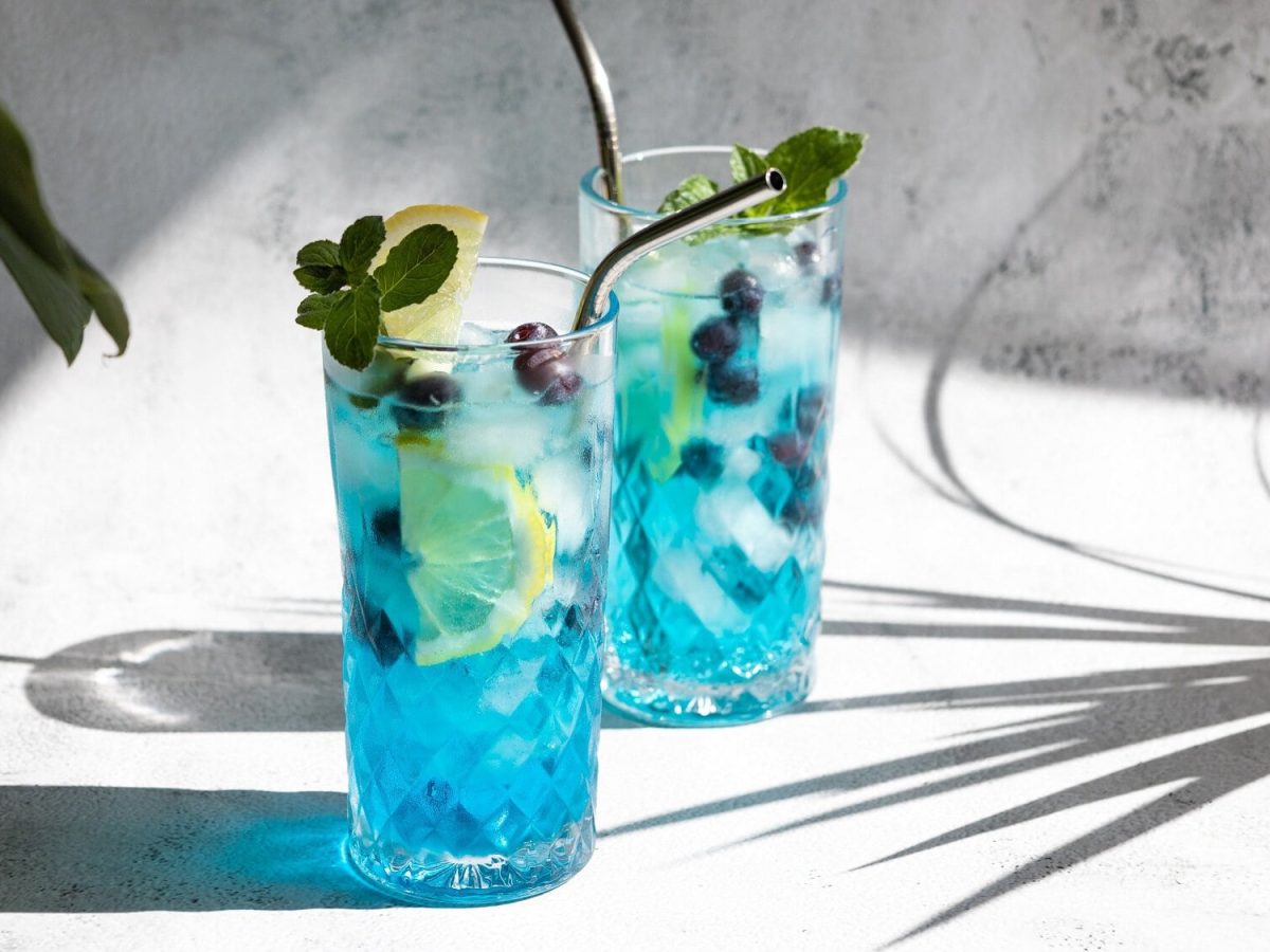 2 Gläser Blue Lagoon mit Eis, Blaubeeren, einer Zitronenscheibe und Metallstrohhalm und Minze garniert in hellem Setting, man sieht Palmenschatten.