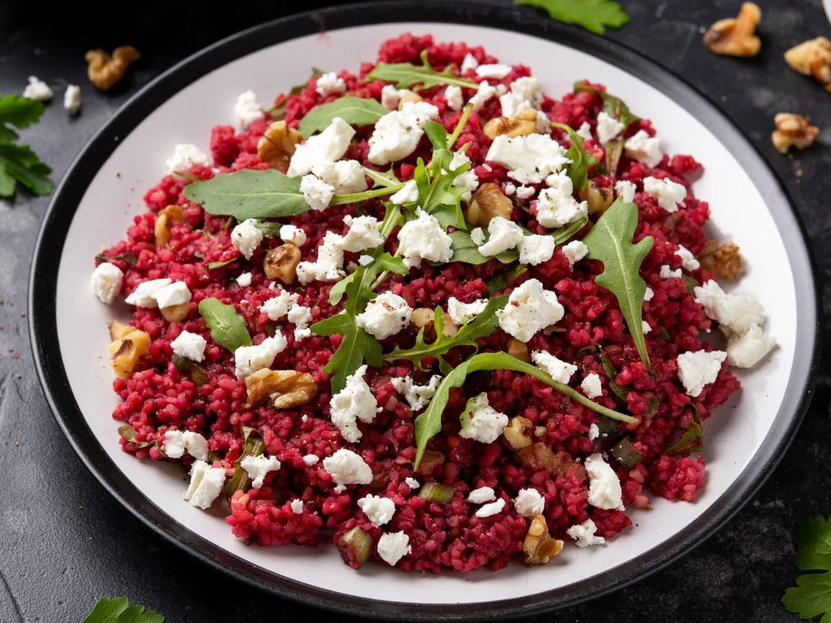 Draufsicht: ein Teller mit Bulgur mit Rote Bete und Walnüssen auf einem dunklen Holztisch. Drum herum sind Walnüsse und Ruccola-Blätter verstreut.