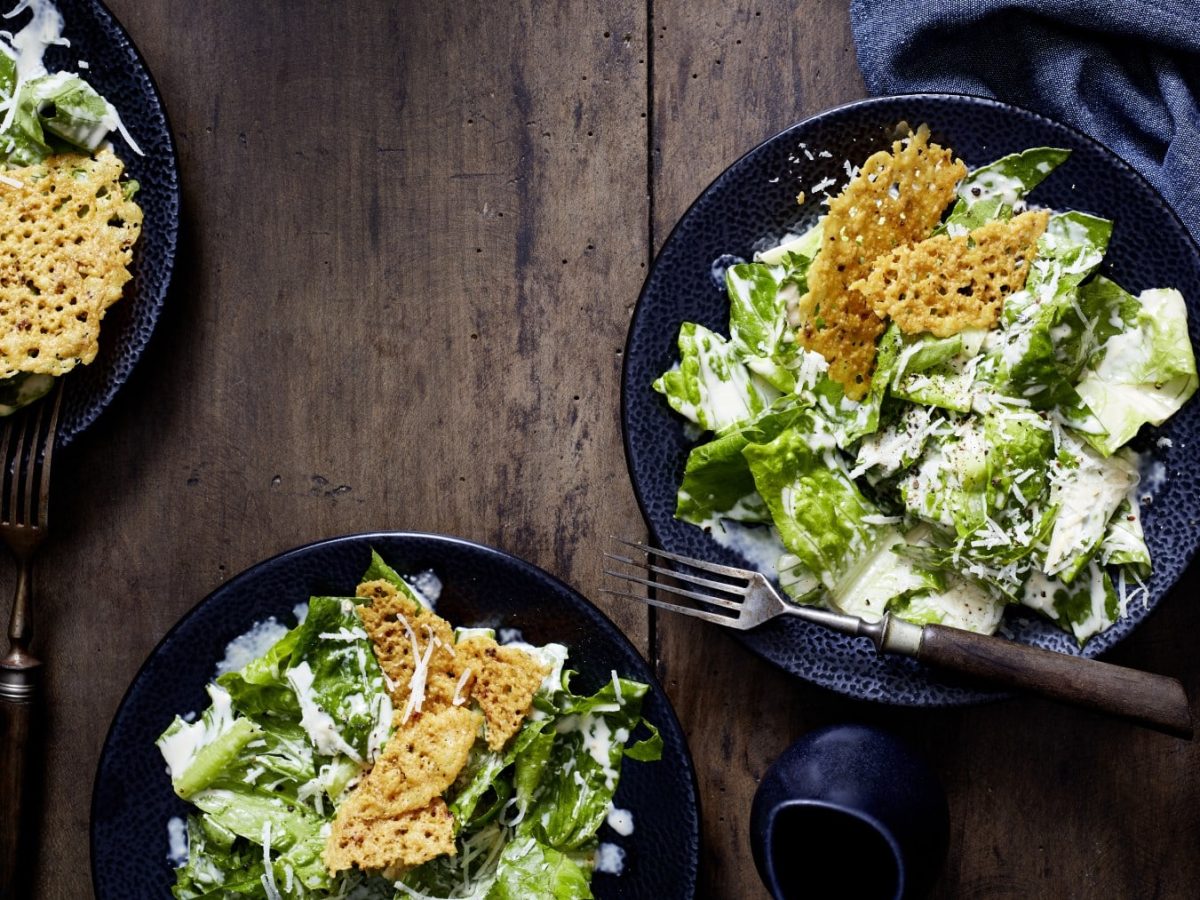 Drei schwarze Teller auf einem hölzernen Hintergrund mit Caesar-Greens und Parmesanbruch und Gabeln. Rechts davon ein schwarzes Geschirrtuch.