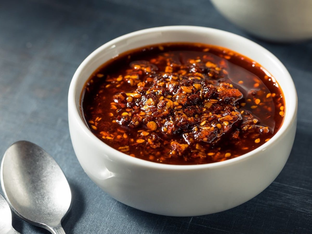 Eine weiße Schüssel mit Garlic-Chili-Crisp auf einem grauen Hintergrund. Links unten zwei Löffel.