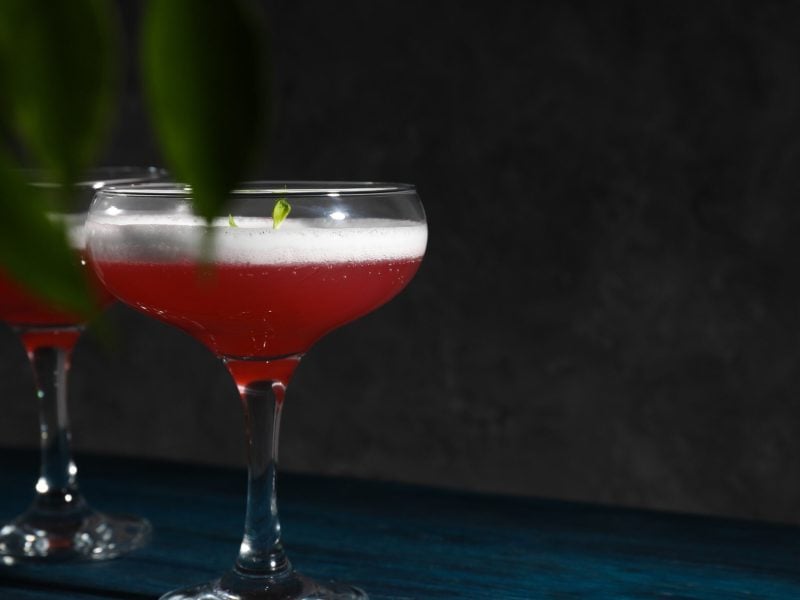 Zwie Cosmo Sour Cocktails in Coupette auf dunklem Untergrund vor dunklem Hintergrund. Im Bild Blätter. Frontalansicht.