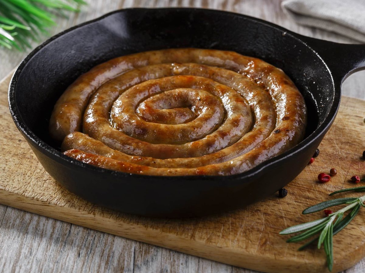 Draufsicht, schräg: eine schwarze gusseiserne Pfanne in der eine gebratene, schneckenförmige, runde Bratwurst liegt. Es handelt sich um eine Englische Wurst-Spezialität. Das ist eine Englische Wurst-Spezialität.