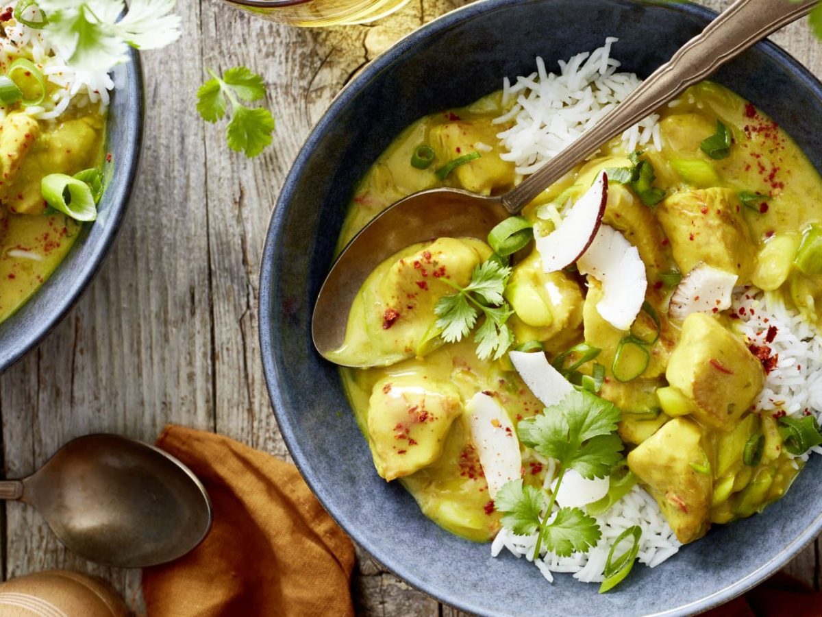 Zwei Portionen Curry-Putengulasch in grauen Schüsseln mit Löffeln auf einem hölzernen Hintergrund. Unten noch ein Löffel, ein oranges Geschirrtuch und eine Pfeffermühle.