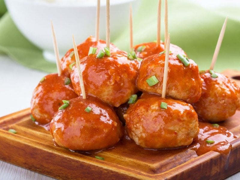 Currywurst-Bällchen mit Holzspießen auf Holzbrett und hellem Untergrund. Frontalansicht.