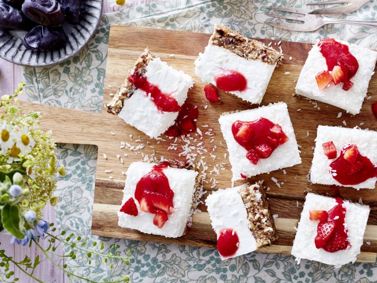 8 Stücke Dattel-Joghurt-Cheesecake auf einem hölzernen Schneidebrett auf einem Geschirrtuch. Rechts unten Honig, links unten Blumen. Links oben eine kleine Schüssel mit Datteln und rechts oben zwei Dessertgabeln.