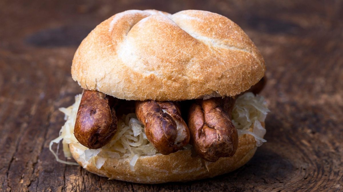 Frontal: Ein aufgeschnittenes Brötchen mit Sauerkraut und drei Bratwürstchen auf dunklem Untergrund. / Drei im Weggla