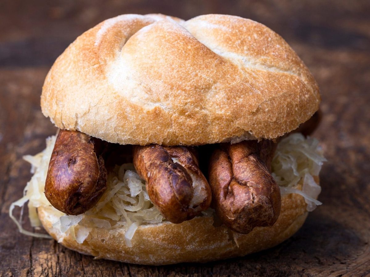 Frontal: Ein aufgeschnittenes Brötchen mit Sauerkraut und drei Bratwürstchen auf dunklem Untergrund. / Drei im Weggla